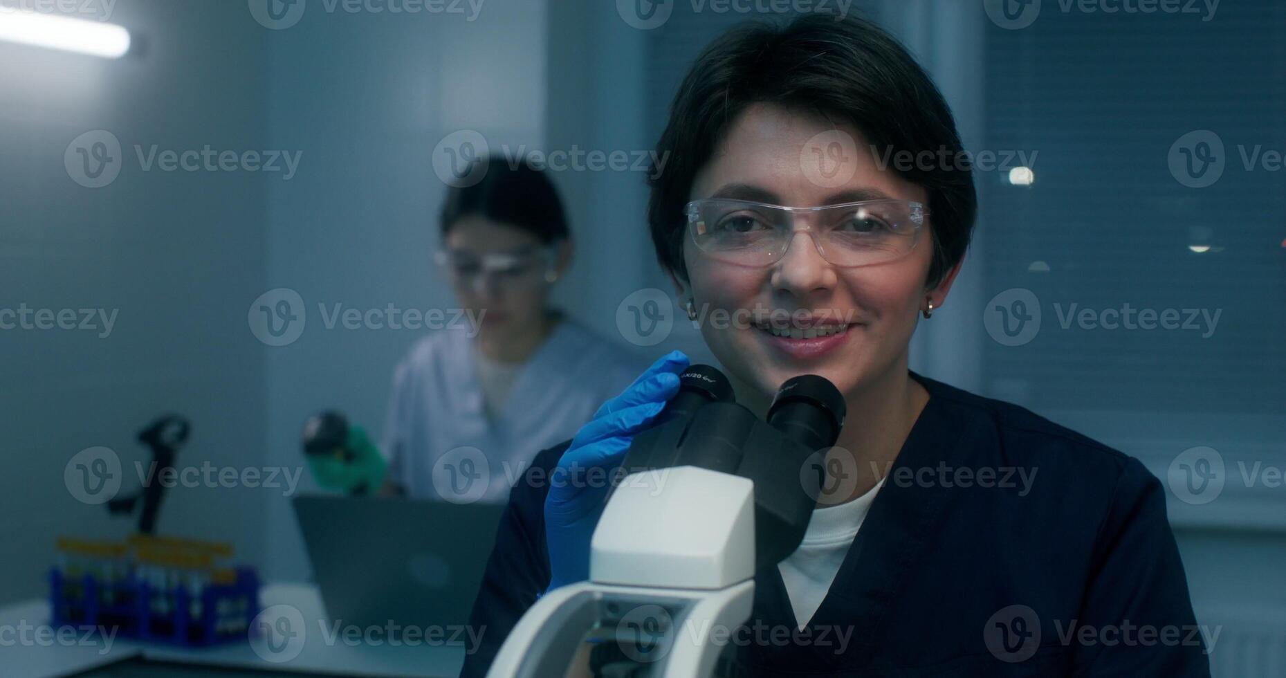 A woman in a lab coat and safety glasses smiles looking at the camera sitting in front of a microscope in a medical laboratory - looking photo
