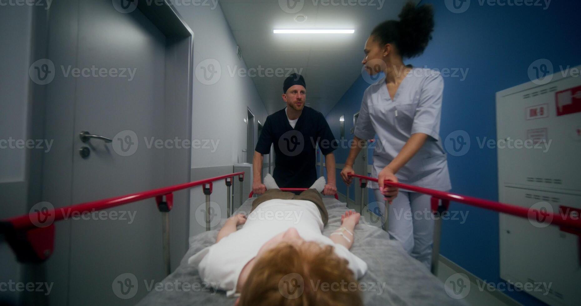 An orderly and a nurse carry a patient on a gurney along the hospital corridor. They walk slowly talking to each other photo