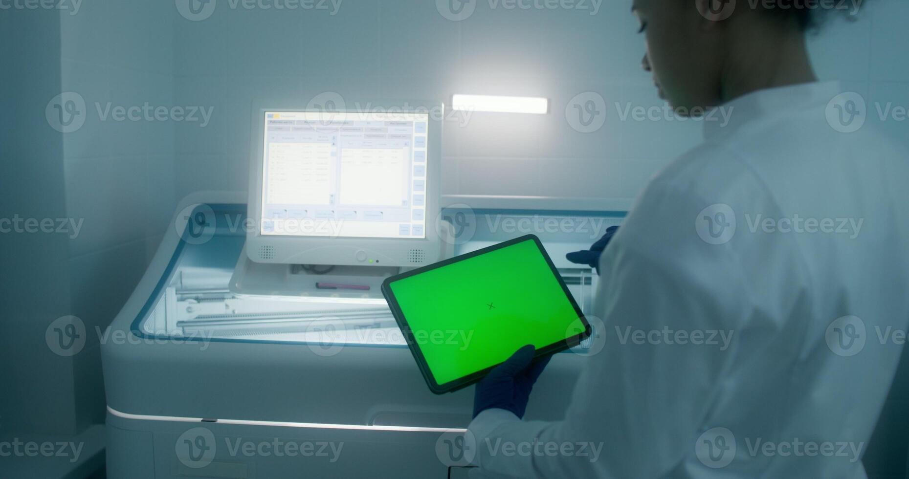 A woman in a lab coat and disposable gloves uses a tablet with a green screen standing in a medical laboratory - medical photo