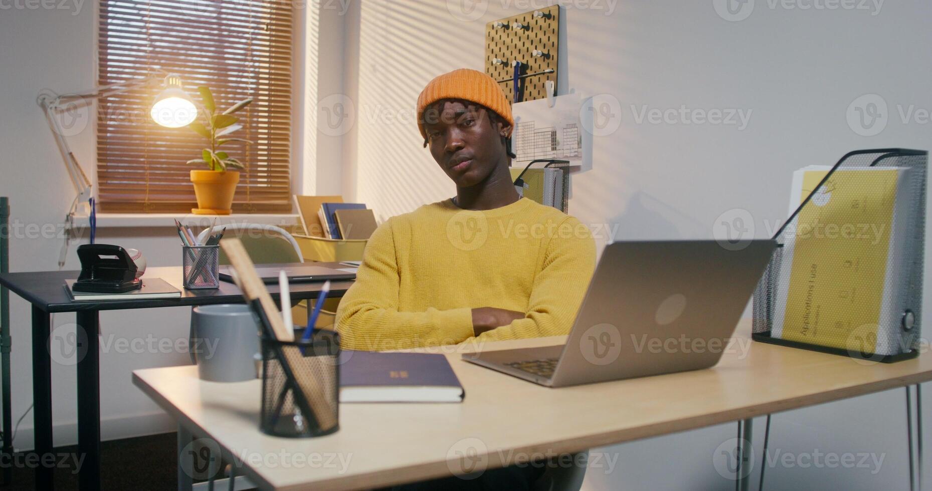 A man in a hat, dressed in casual clothes, looks at the camera in a serious manner while sitting in front of an open laptop in the office - camera photo