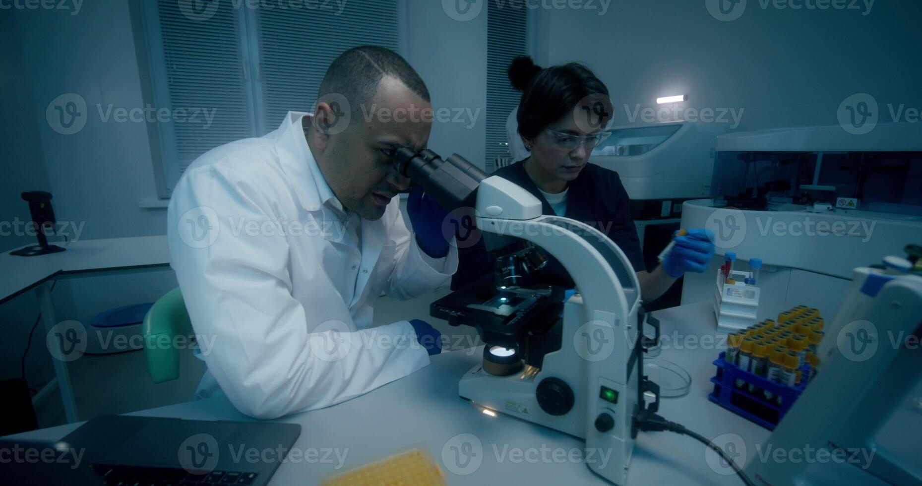 A man and a woman in lab coats are working in a medical laboratory, conducting a blood test and using a microscope. They are talking sitting at the table photo