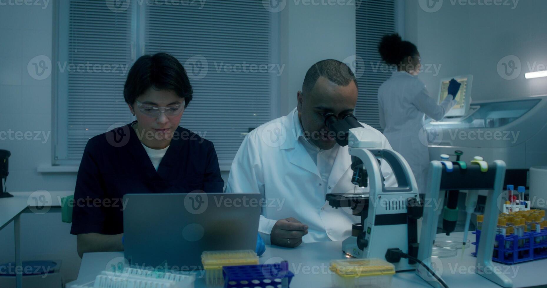 Work in a medical laboratory. A man uses a microscope to perform a blood test. Woman typing on laptop sitting next to him photo