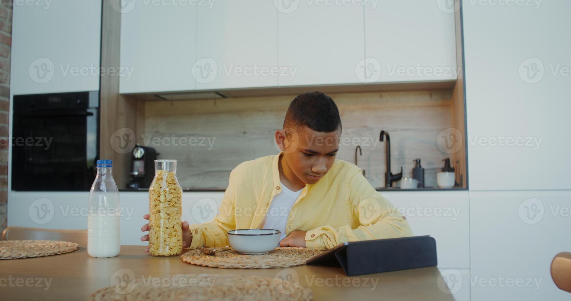 The boy has breakfast at home, he pours cereal into a plate while sitting at the table and looking at the tablet - sitting photo
