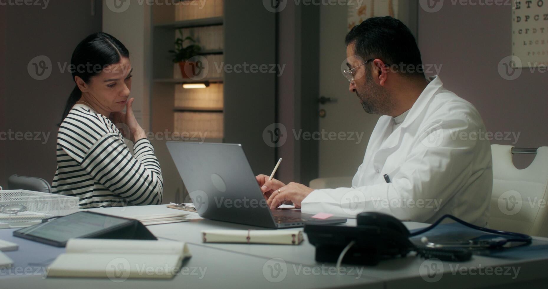 The doctor listens to the patient's complaints sitting in front of an open laptop at the table and writes in a notebook. Medical appointment at the clinic photo