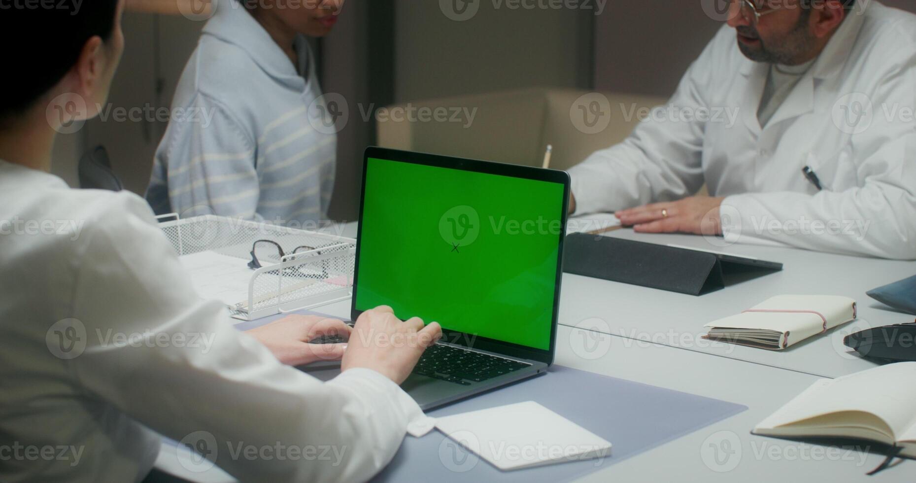 The nurse uses a laptop with a green screen, entering patient data. A woman talks to a doctor sitting at a table in the background - doctor photo
