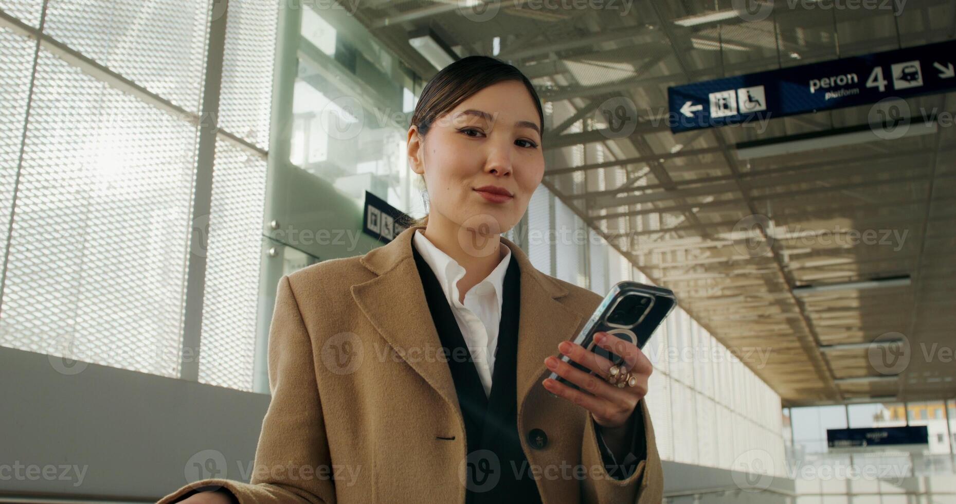 A woman in a coat smiles looking at the camera standing in the railway station crossing. Her mobile phone and luggage are in her hand photo