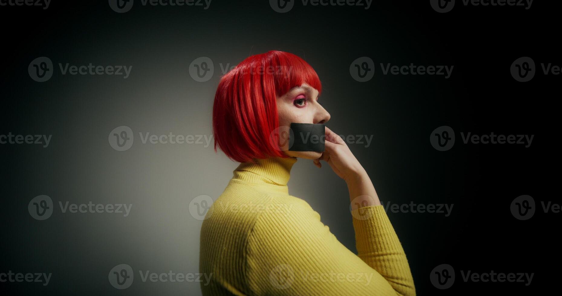 A woman in an orange wig rips the tape from her mouth and looks at the camera, standing on a gray background in the studio photo