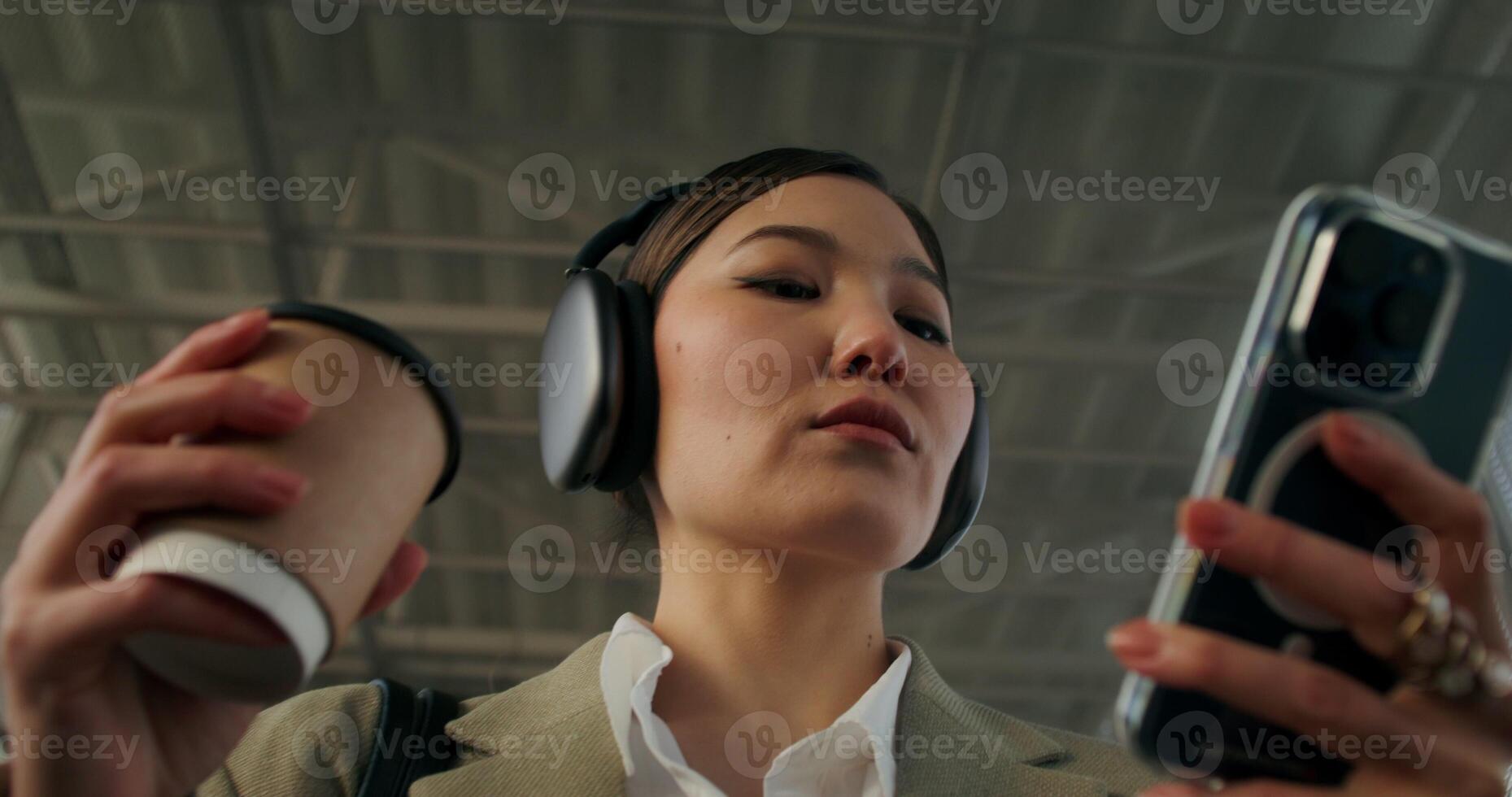 A woman in formal clothes and with headphones on her head uses a mobile phone and drinks coffee from a disposable cup walking along the railway elevated passage - coffee photo