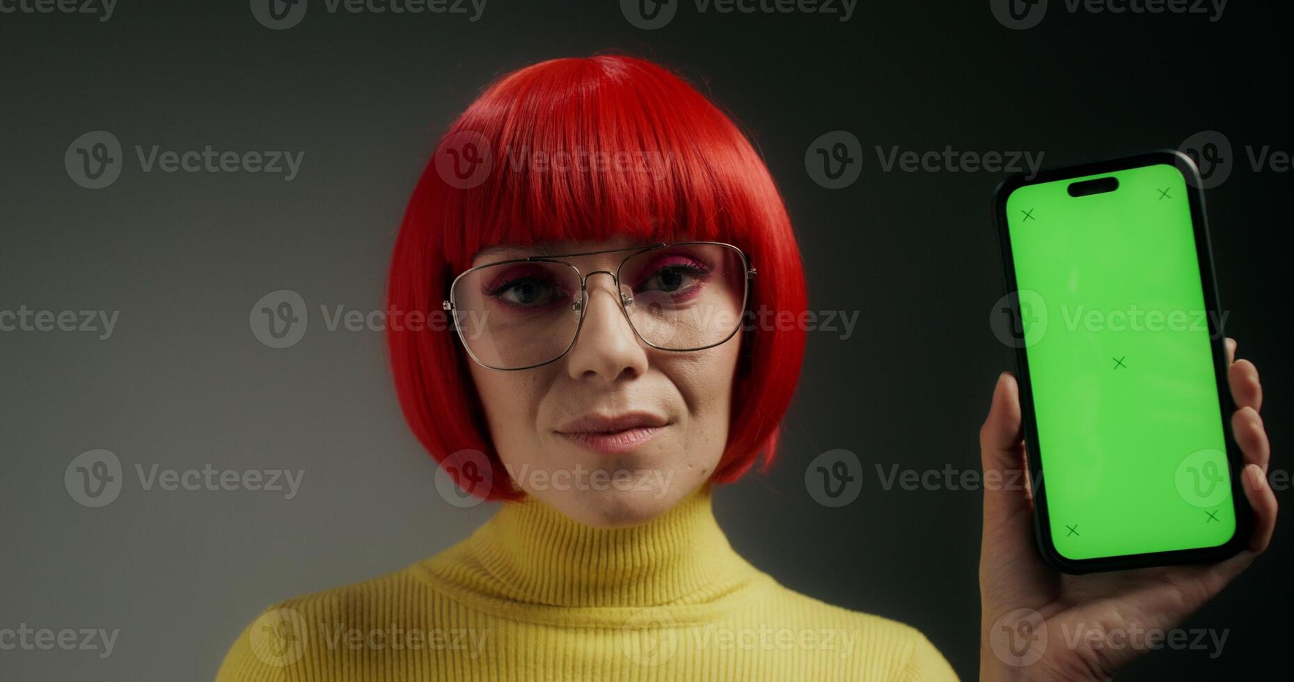 A woman with glasses and an orange wig smiles at the camera, holding a mobile phone with green screen in her hand, standing on a gray background in the studio - smiles photo