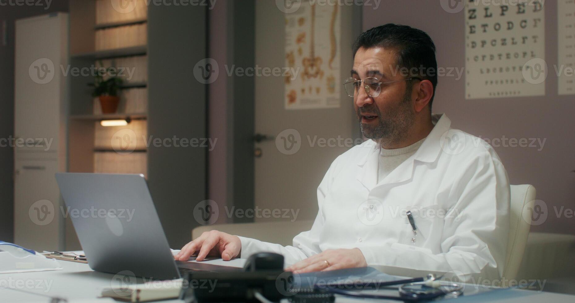 The doctor is talking on a chat on a laptop sitting at a table in the medical office at the clinic - medical photo