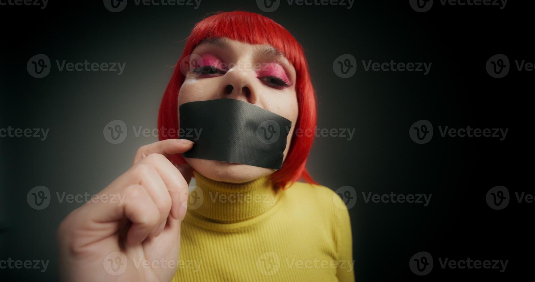 A woman in an orange wig rips the tape from her mouth and looks at the camera, standing on a gray background in the studio - background photo