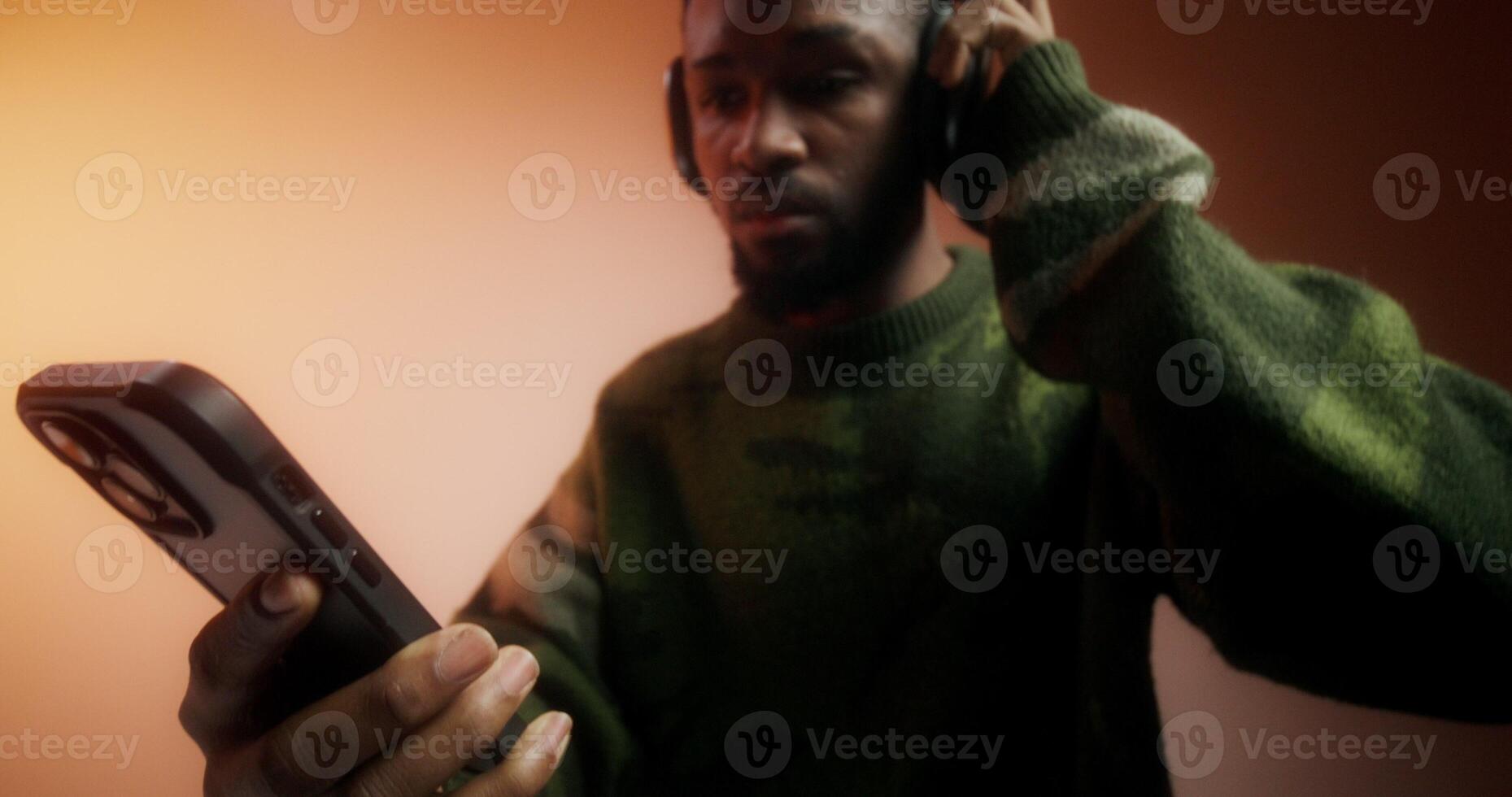 A bearded man in a sweater and headphones turns on music on his mobile phone and starts dancing standing on an orange background in the studio - headphones photo