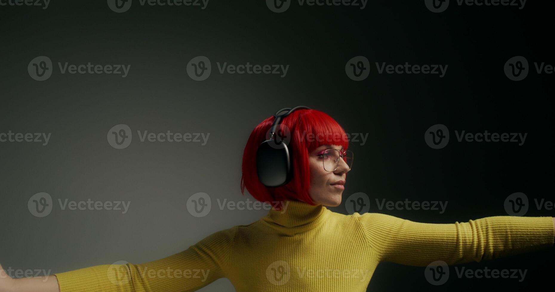 A woman wearing glasses, an orange wig and headphones, dancing with her hands moving while standing on a gray background in the studio photo