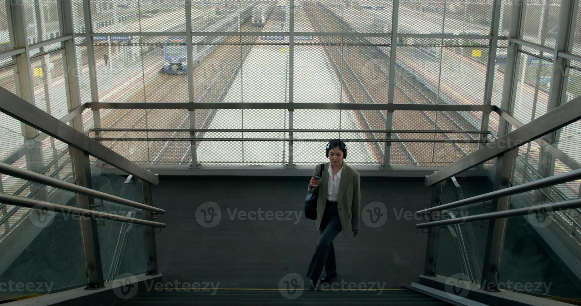 A woman in formal clothes and headphones walks up the stairs in the elevated passage of the railway station and drinks coffee from a disposable cup - headphones photo