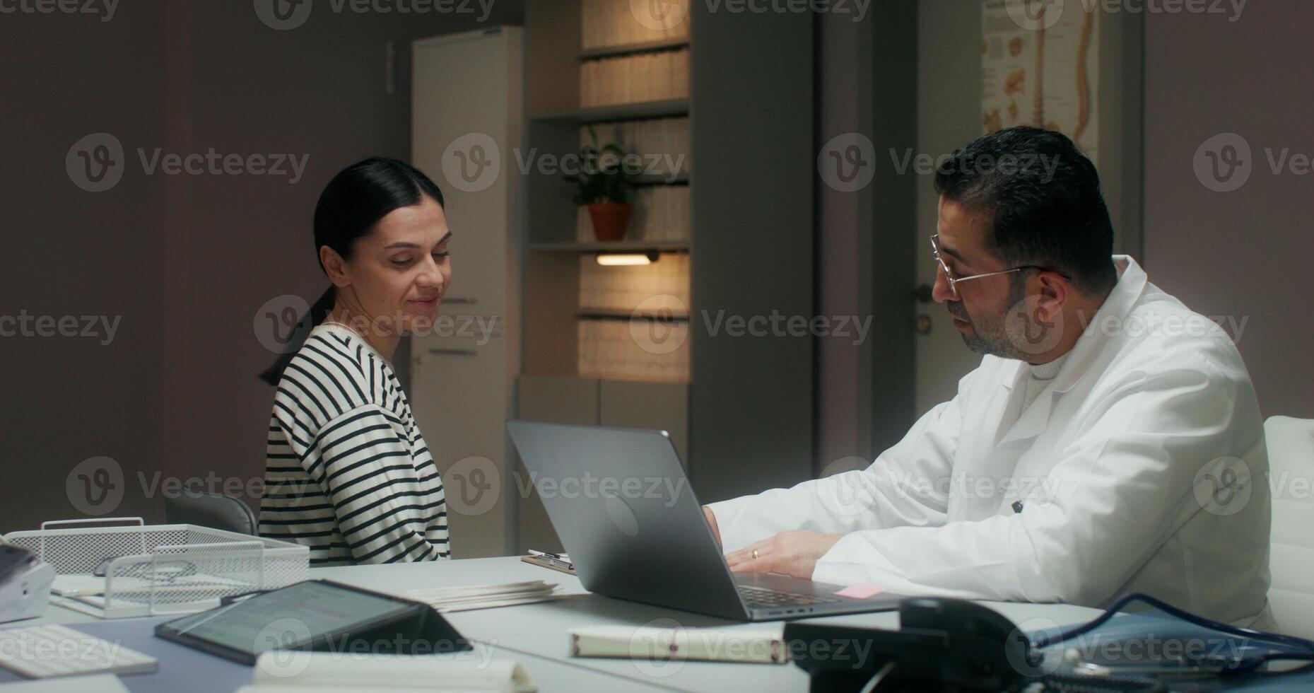 A female patient and a doctor smile and talk sitting in a medical office. A man writes in a notebook, making an appointment for treatment photo
