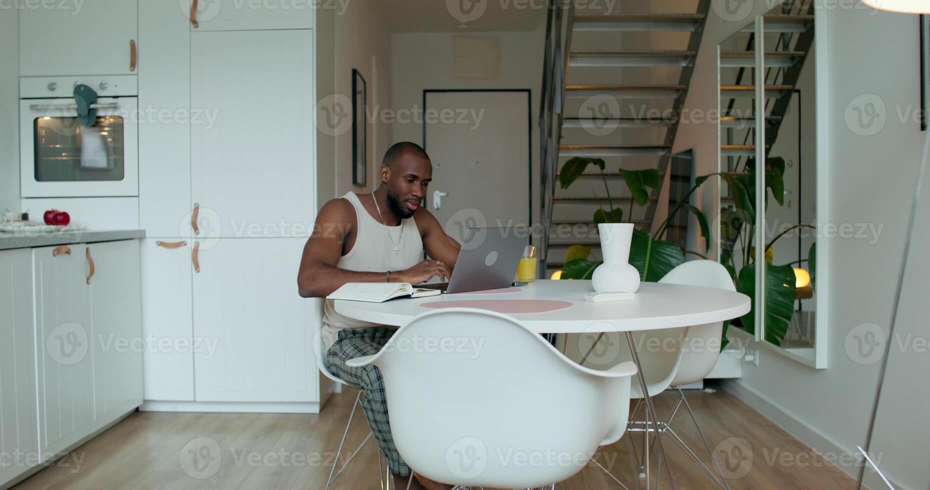 A man in pajamas uses a laptop and writes in a notebook sitting at a table in the kitchen at home with a serious look photo