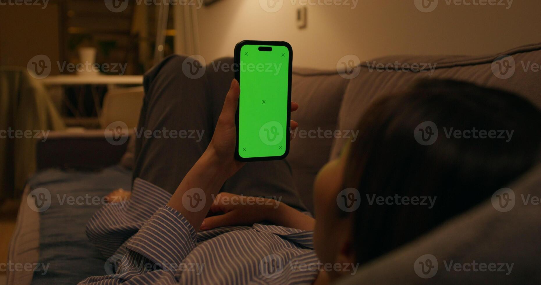 A woman uses a mobile phone with a green screen lying on the couch at home, dimmed evening light - dimmed - uses photo