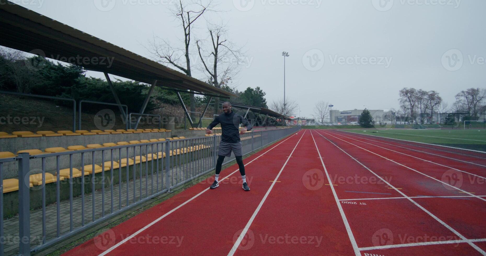 A man in a sports outfit with earphones in his ears does a warm-up standing at the stadium on a cold day - standing - in photo