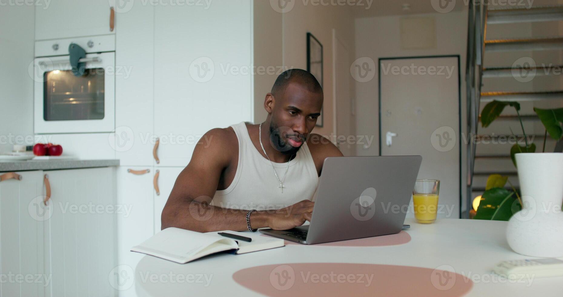 A man in pajamas uses a laptop and writes in a notebook sitting at a table in the kitchen at home with a serious look - writes photo