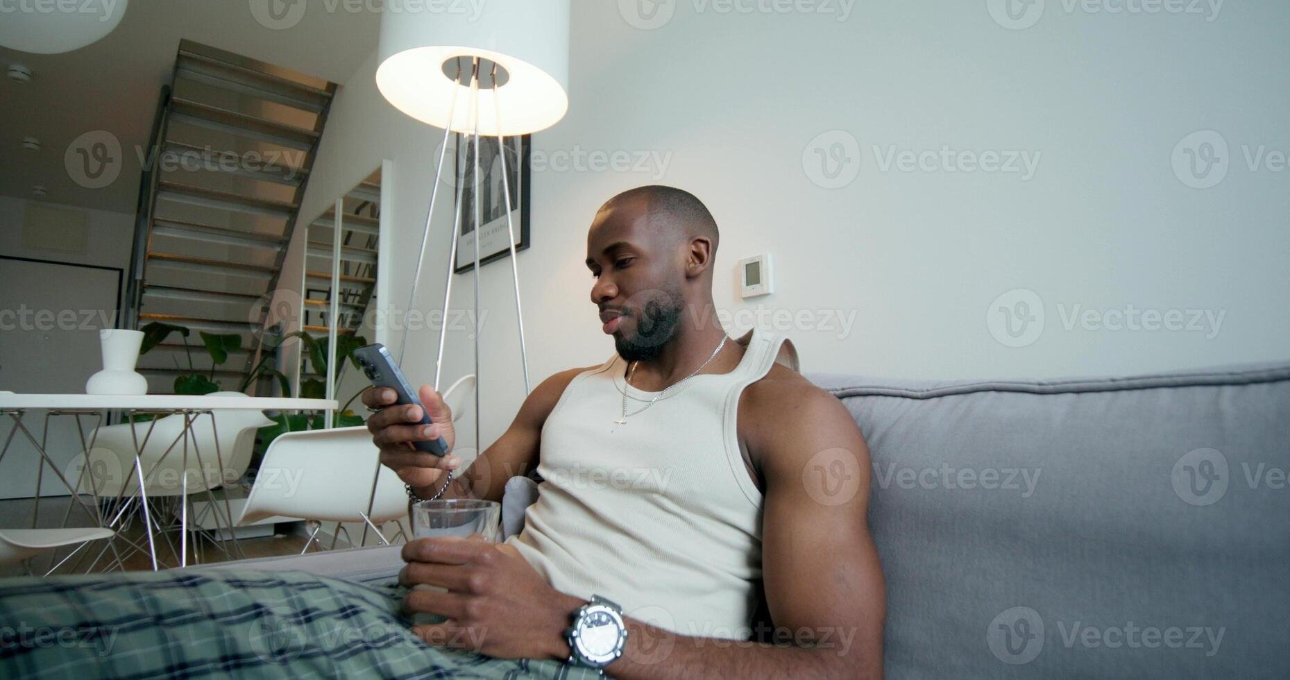 A man in pajamas uses a mobile phone and drinks water from a glass sitting on the couch at home. Daylight - drinks photo