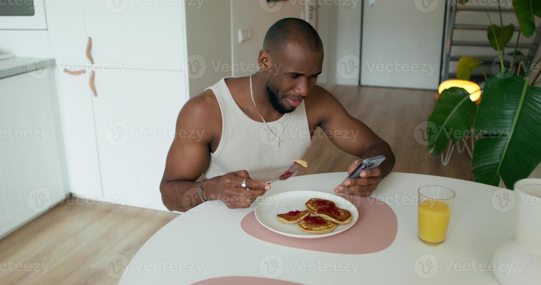 A man in pajamas eats pancakes and uses a mobile phone sitting at a table in the kitchen at home. Daylight - sitting photo