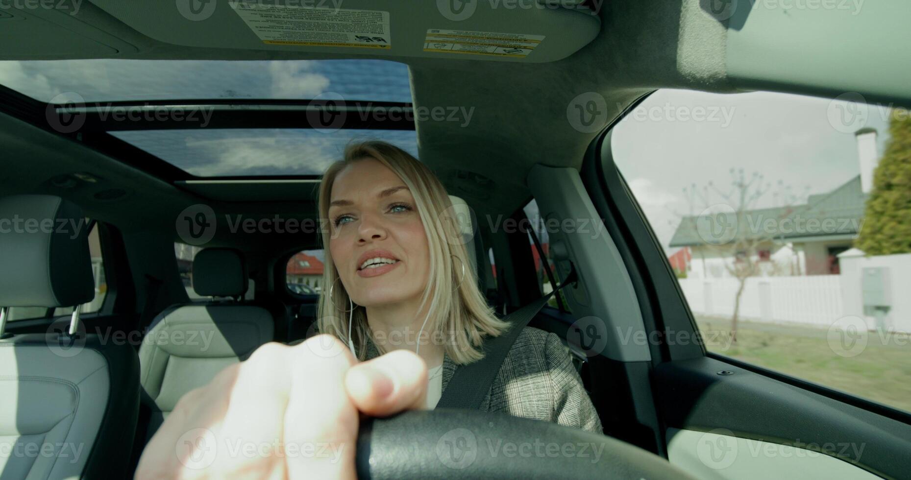 A stylish woman with earphones is driving around the city on a sunny day. She is smiling talking on the phone - driving photo