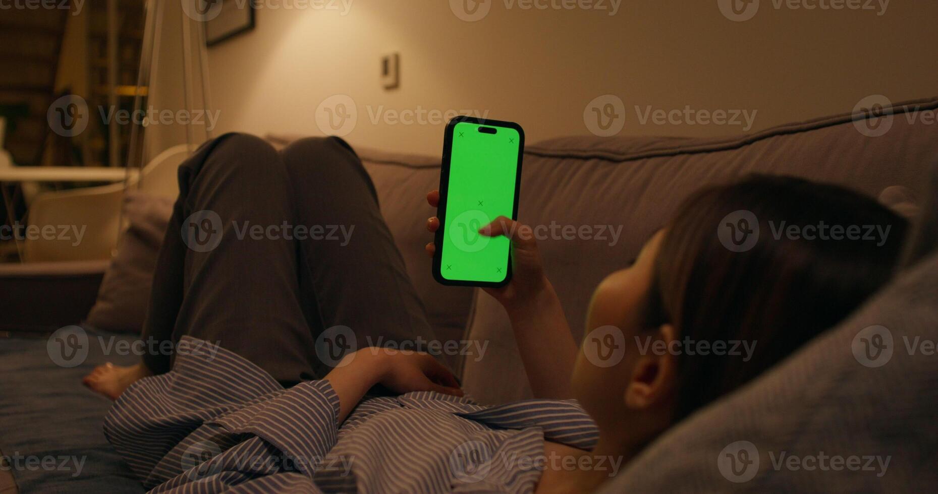 A woman uses a mobile phone with a green screen lying on the couch at home, an unrecognizable person photo