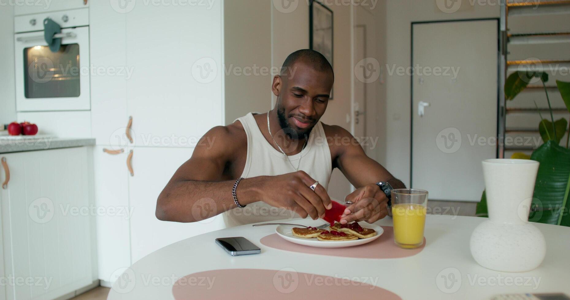 A man in pajamas squeezes jam out of a package onto pancakes while having breakfast, sitting at the table at home. Daylight - package photo