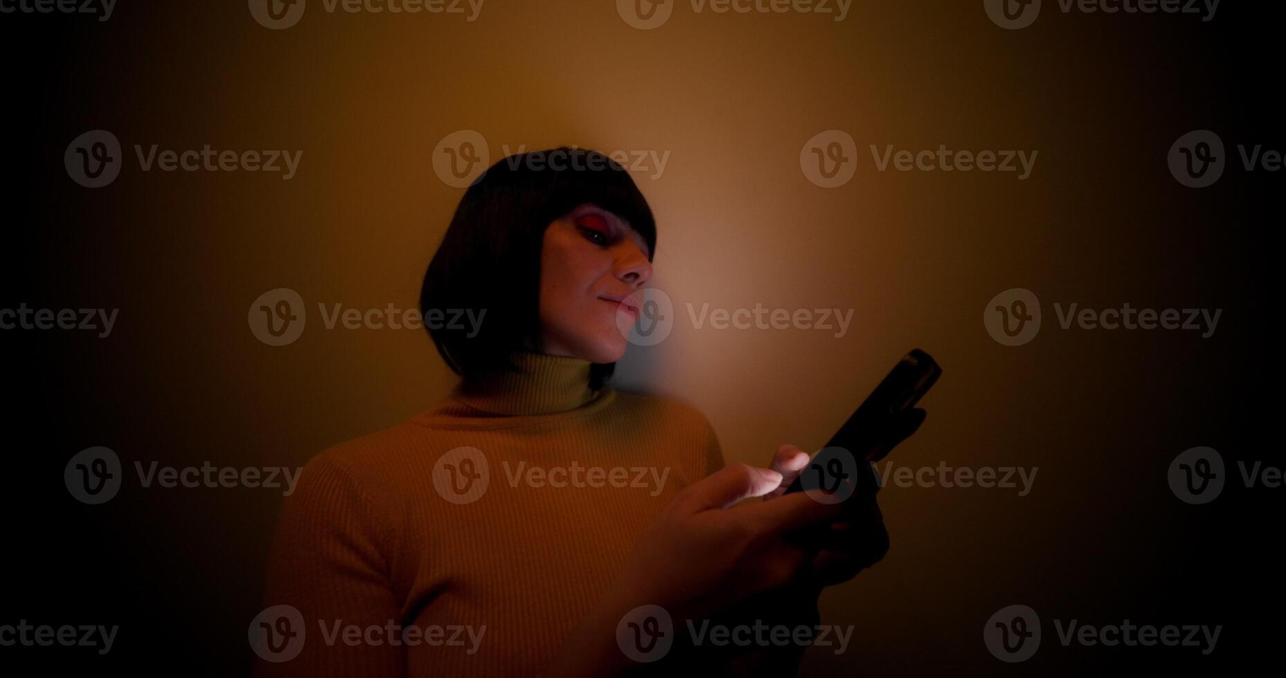 A woman in a wig uses a mobile phone, leaning against the wall in the orange backlight in the studio photo