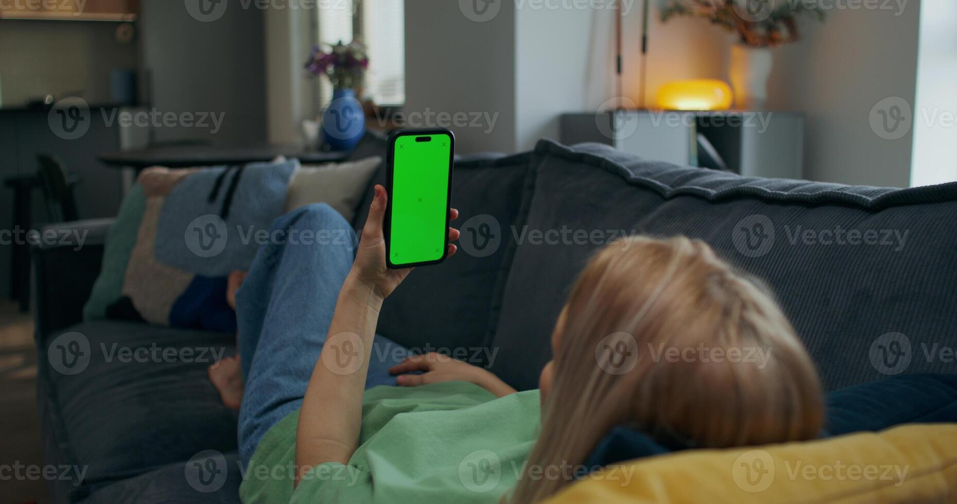 A woman holds a mobile phone with green screen in her hand. She is lying on the couch at home, rear view, unrecognizable person photo