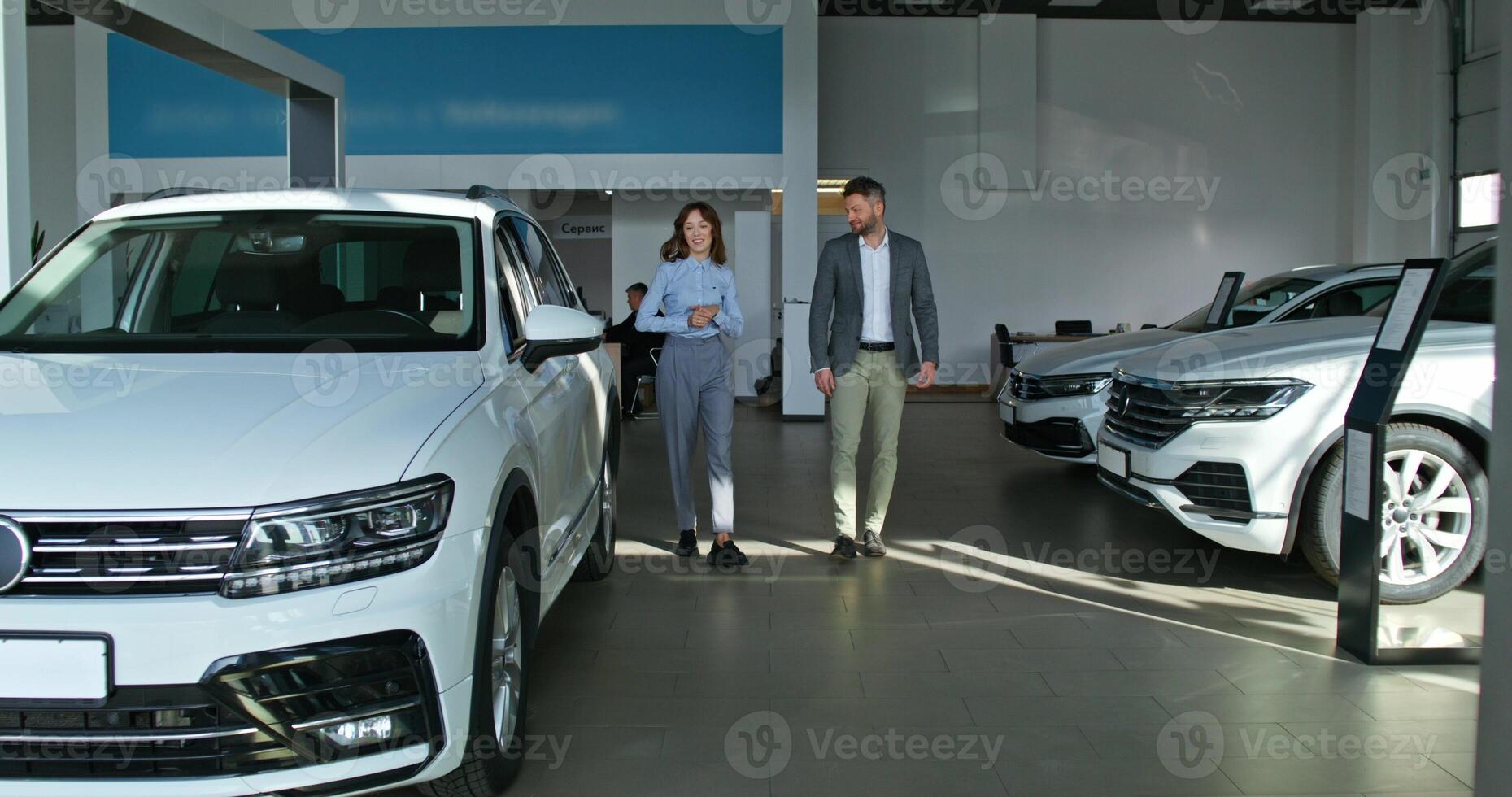 A car dealership manager in formal clothes talks to a customer and shows him a car standing in the showroom - clothes photo