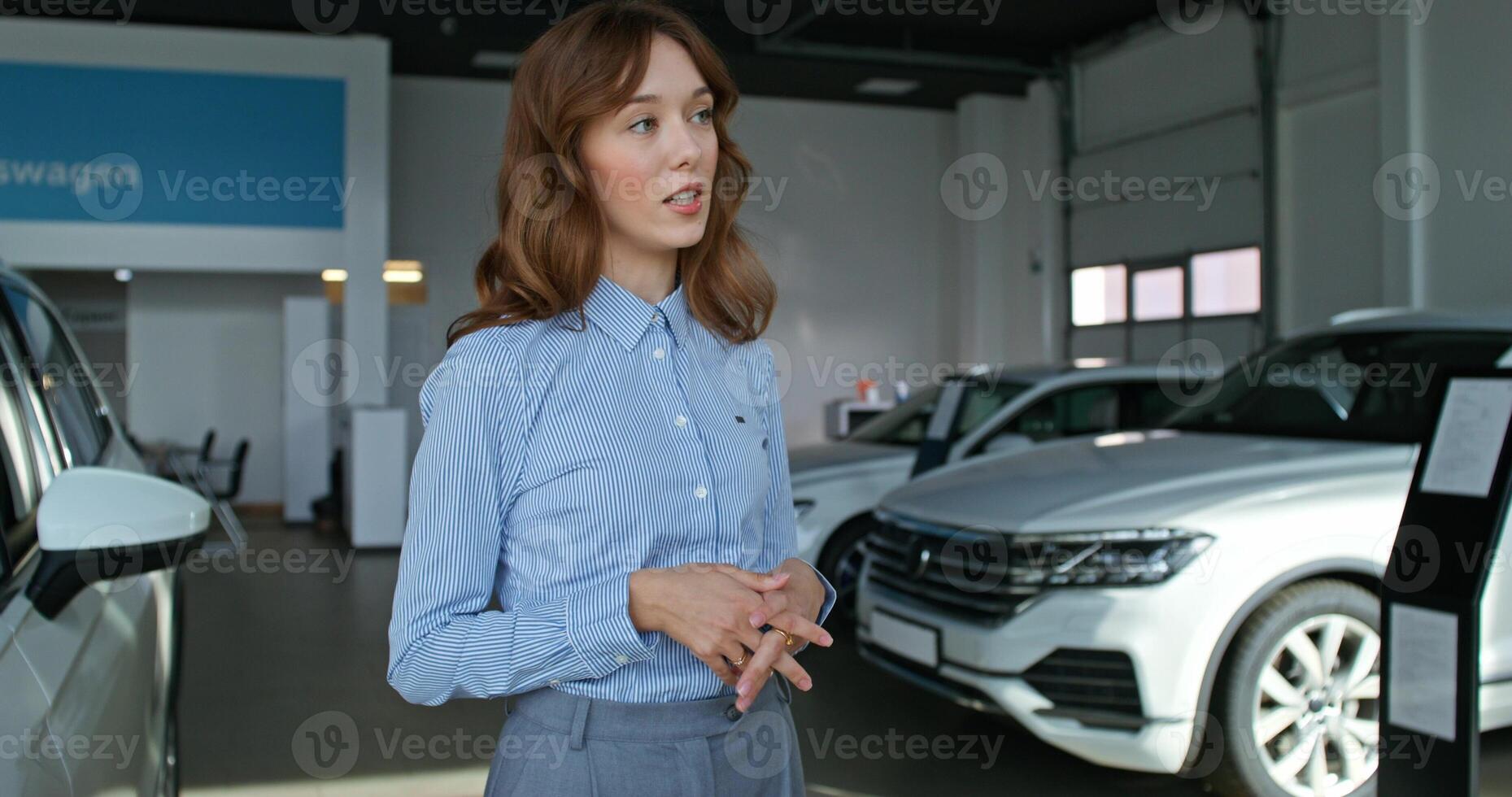 A car dealership manager in formal clothes smiles and talks to a customer standing near cars in a car dealership photo