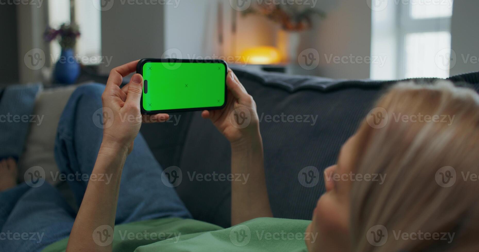 A woman holds a mobile phone with green screen in her hand. She is lying on the couch at home, unrecognizable person - unrecognizable photo