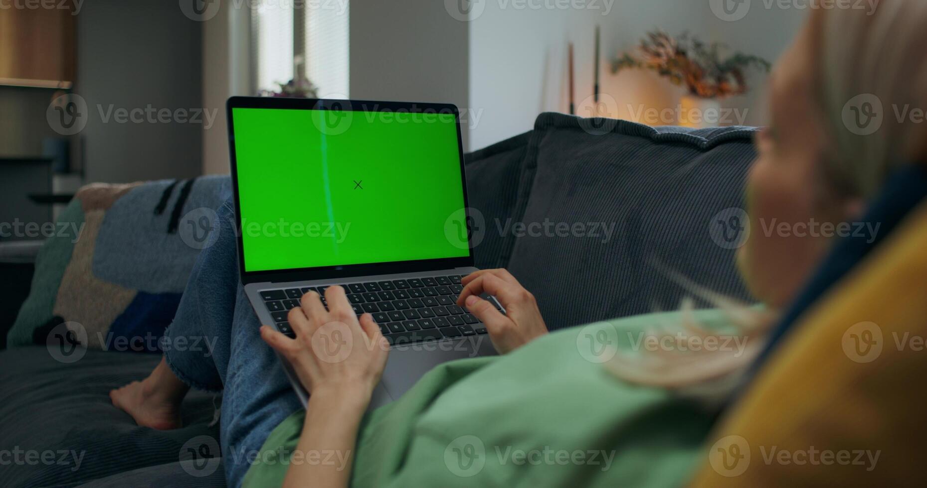 A woman uses a laptop with green screen lying on the couch at home, an unrecognizable person - unrecognizable photo