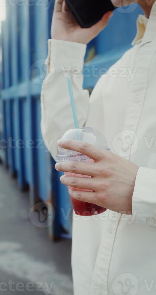 A young man of Chinese appearance is talking on a mobile phone, and drinking juice from a disposable cup with a straw standing near a metal wall outdoors - mobile photo