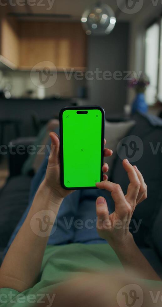 A woman uses a mobile phone with green screen lying on the couch at home. A rear view of her hands, an unrecognizable person photo