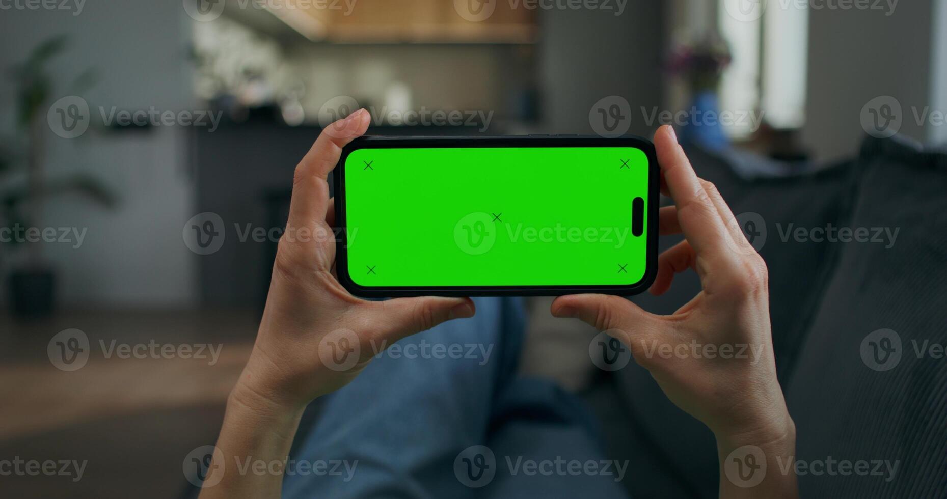 A woman holds a mobile phone with green screen in her hand. She is lying on the couch at home, close-up of her hands, unrecognizable person photo