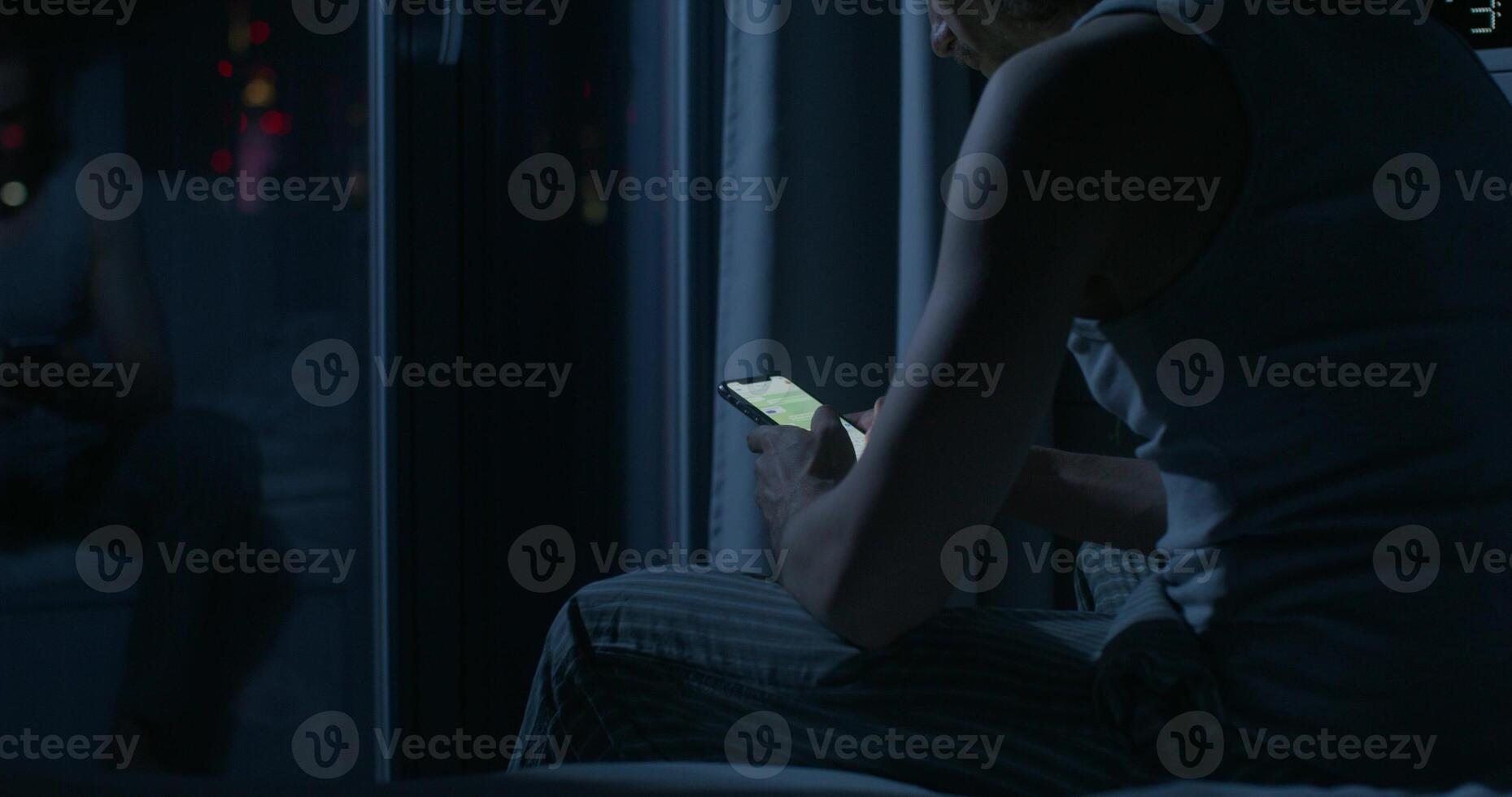 A man uses a mobile phone while sitting on the bed at night. Close-up of his hands, an unrecognizable person. Dark room, home interior photo