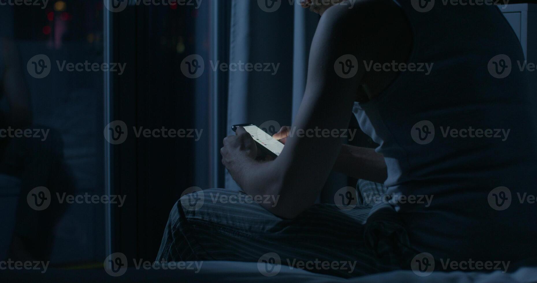 A man uses a mobile phone while sitting on the bed at night. Close-up of his hands, an unrecognizable person. Dark room, home interior - unrecognizable photo