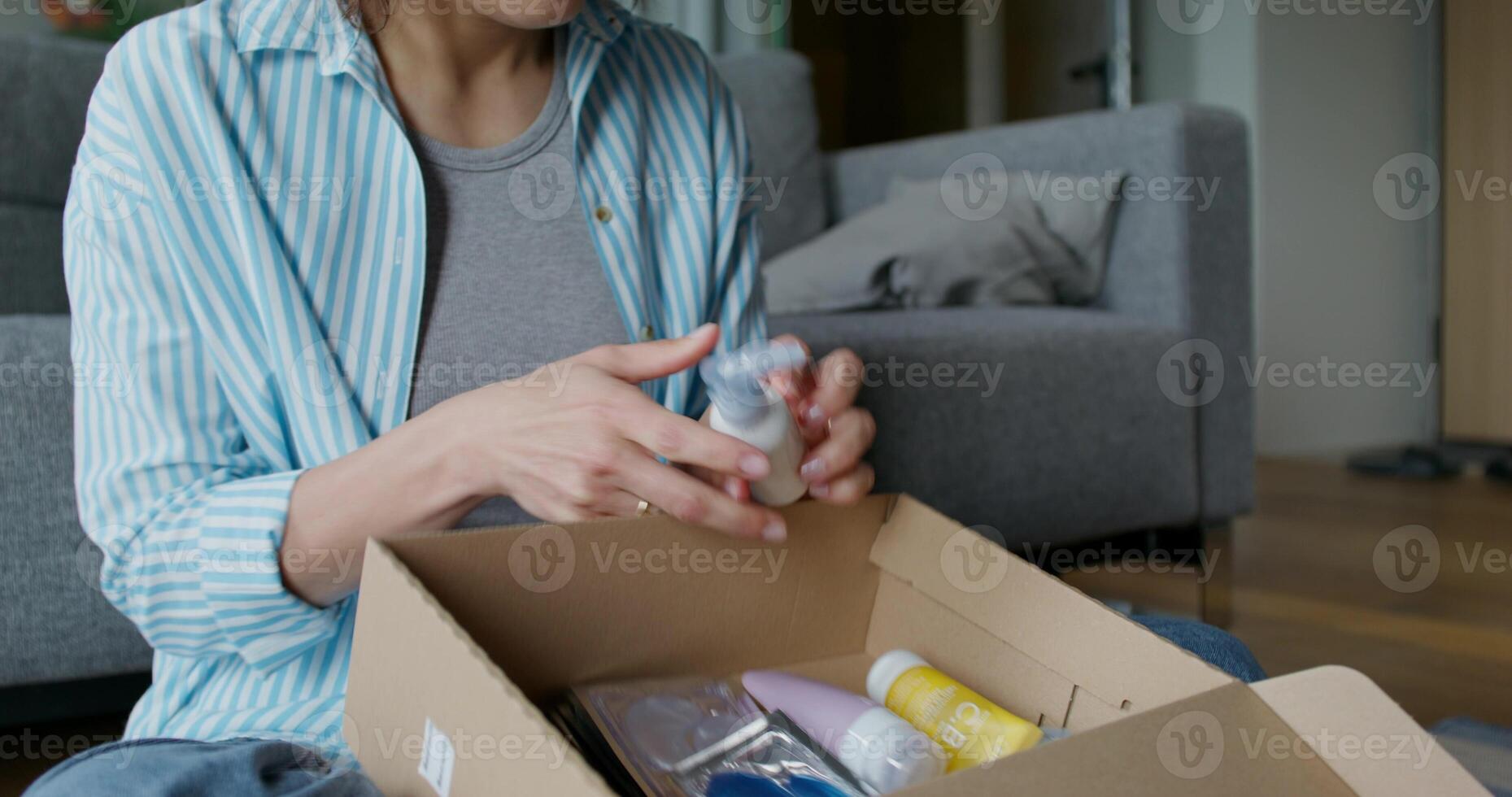 The woman opens the box and pulls out the cosmetics, she smiles. She is sitting on the floor near the sofa at home. Daylight - sitting photo