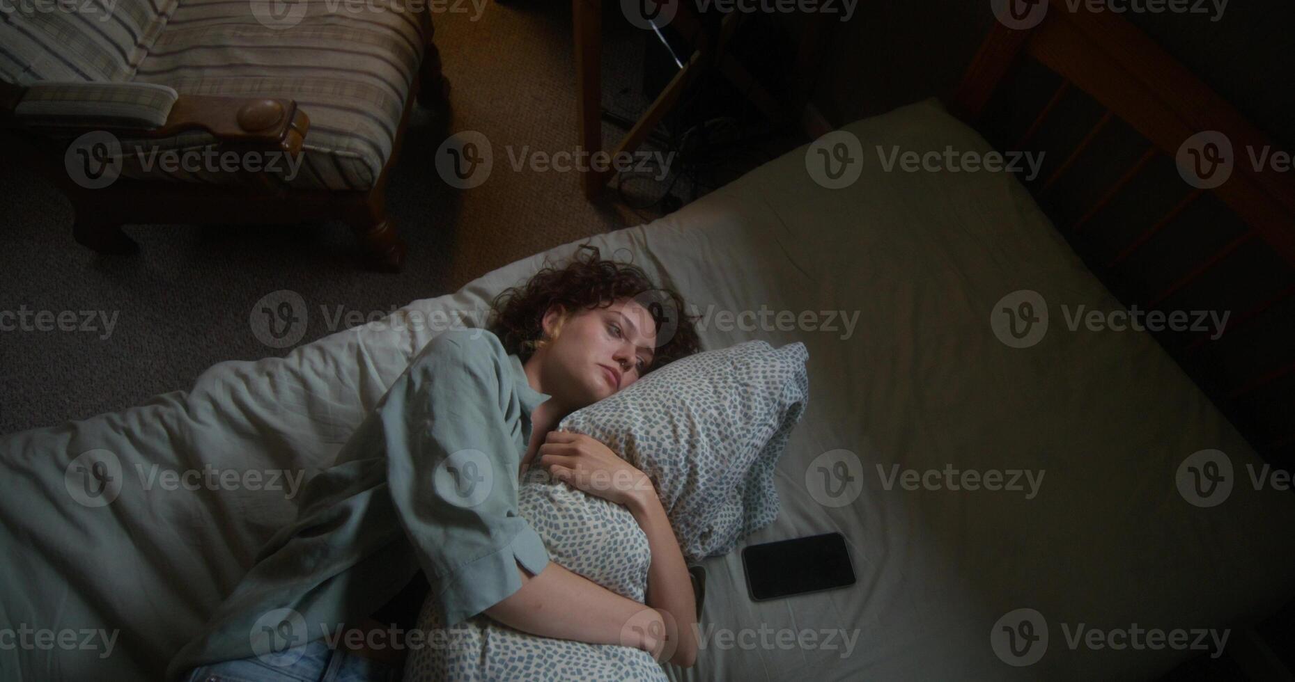 A woman is crying and hugging a pillow lying in bed at home. The mascara is smeared under the eyes. The mobile phone is on her bed photo