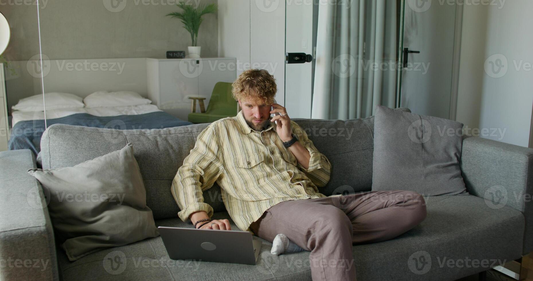 A bearded man uses a laptop and talks on a mobile phone sitting on the couch at home with a serious look. Daylight photo