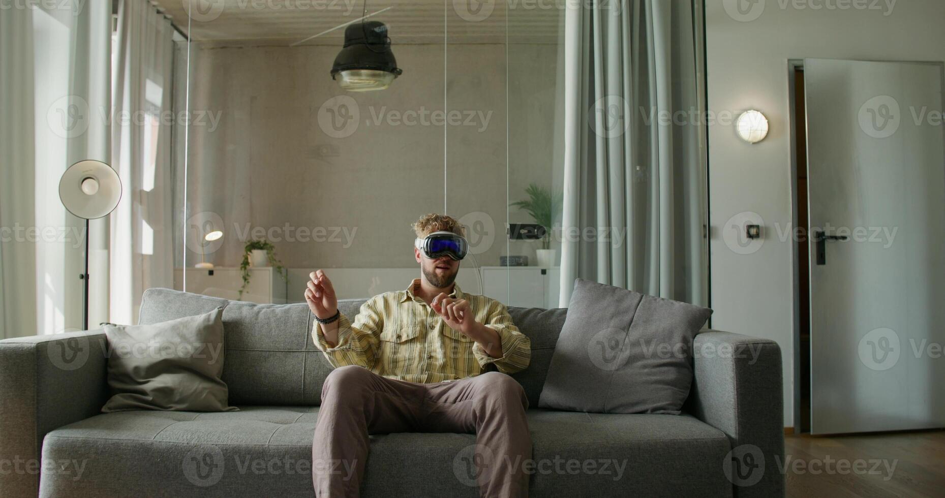 A man in a virtual reality headset moves his hands while sitting on a sofa in a home interior, daylight photo