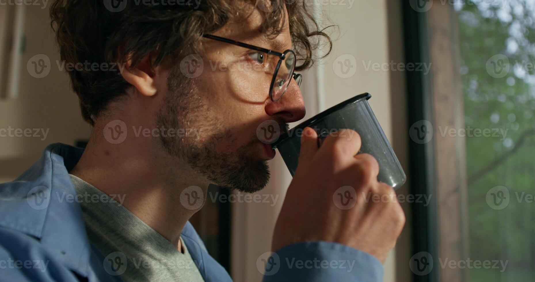 A curly-haired man with glasses drinks coffee with a thoughtful look standing at home photo