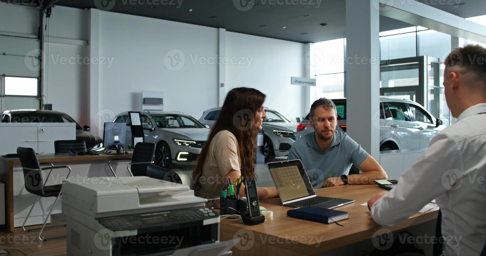 The car dealership manager uses a laptop when making a car purchase. A man and a woman are smiling sitting at the table opposite the manager photo
