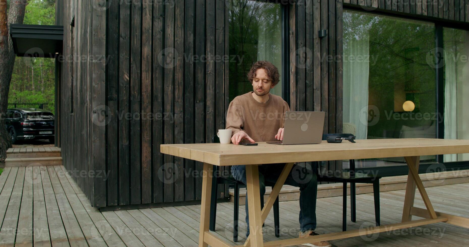 A man in casual clothes uses a laptop sitting at a table on the terrace of a stylish cottage on a summer day. He's answering his mobile phone. - laptop photo