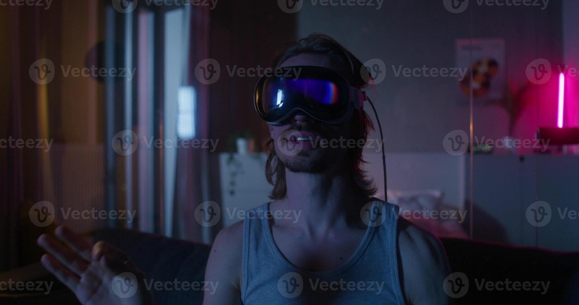 A man wearing virtual reality glasses is sitting on the couch and moving his arms. Dark room, home interior - reality photo
