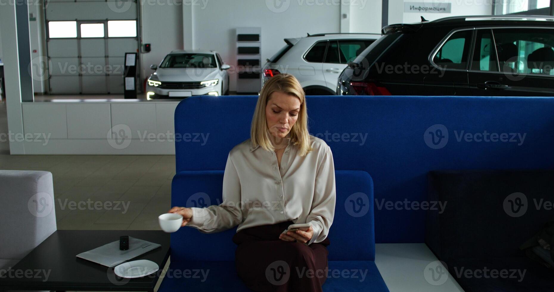 A stylish woman uses a mobile phone and drinks coffee sitting on the couch in a car dealership photo