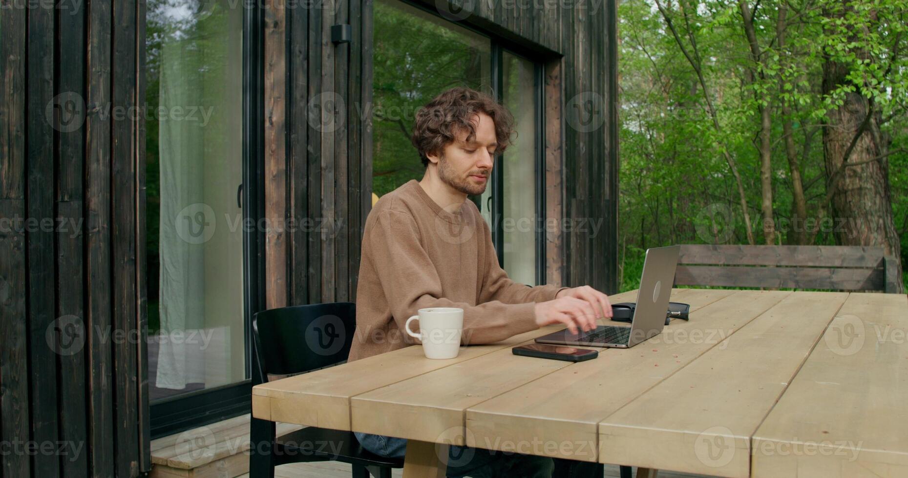 A man in casual clothes uses a laptop sitting at a table on the terrace of a stylish cottage on a summer day. He's answering his mobile phone. - terrace photo