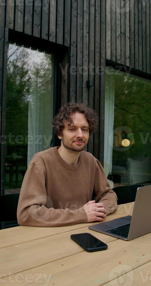 A man in casual clothes smiles looking at the camera sitting in front of an open laptop on the terrace of a stylish cottage on a summer day - smiles photo